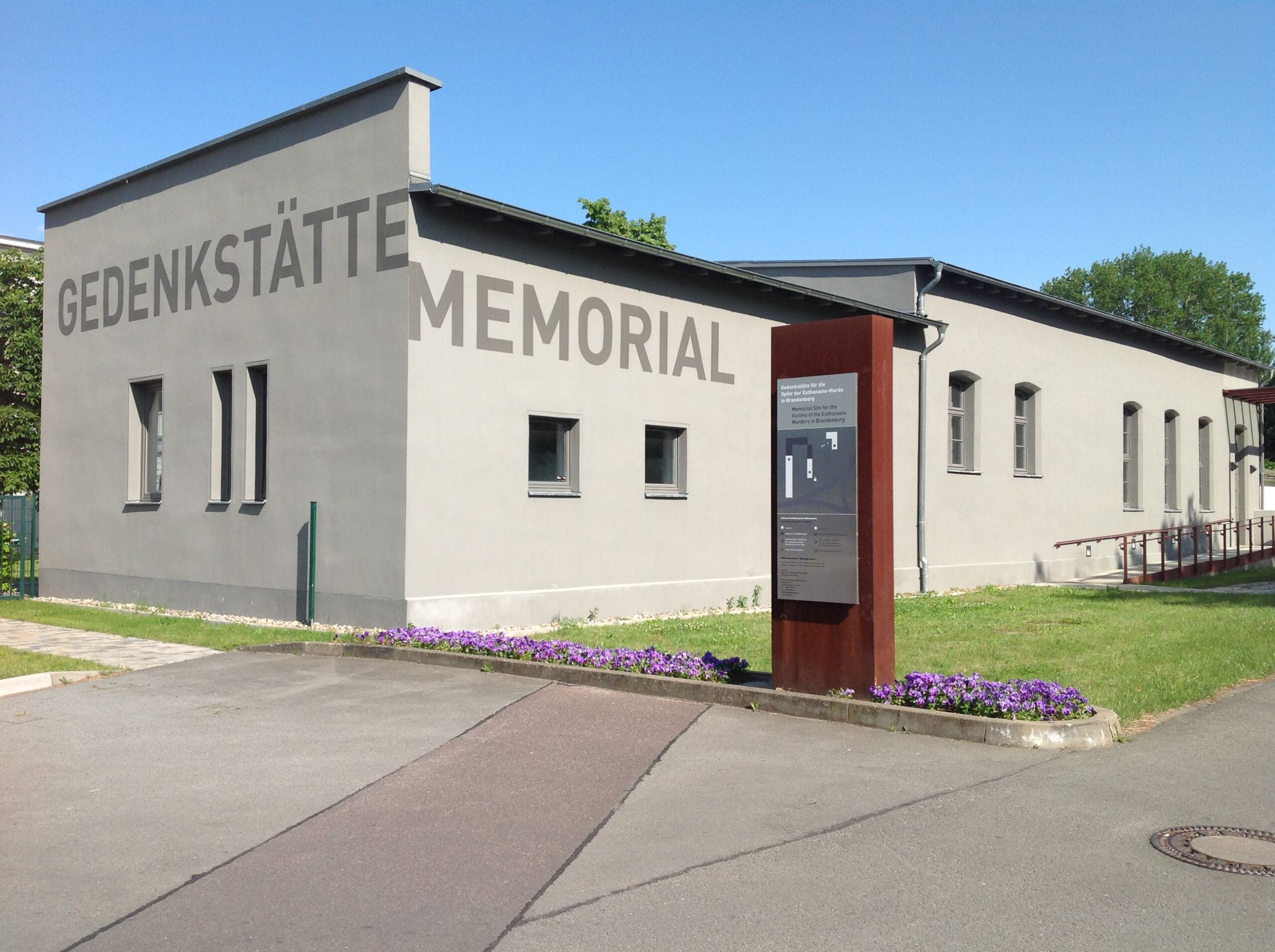 Die Gedenkstätte von außen. Am Gebäude steht Gedenkstätte und Memorial. Davor eine Informationstafel. Das Foto ist in Farbe.